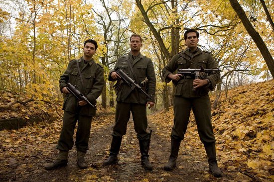 Drie mannen in Duitse militaire uniformen staan in een bos met herfstbladeren, elk met een machinepistool.