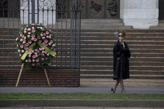 Een vrouw in een zwarte trenchcoat rookt bij een grote krans met roze bloemen.