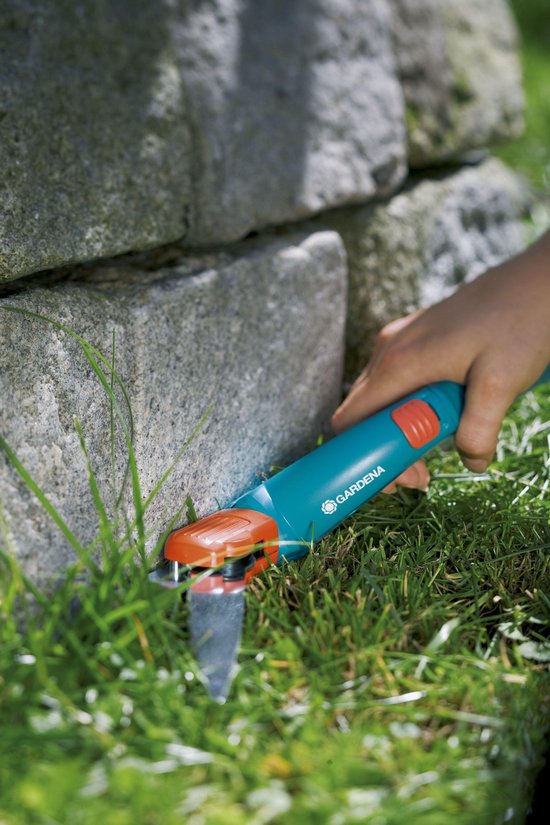 Hand maait gras langs stenen muur met blauwe Gardena graschaar met oranje details.
