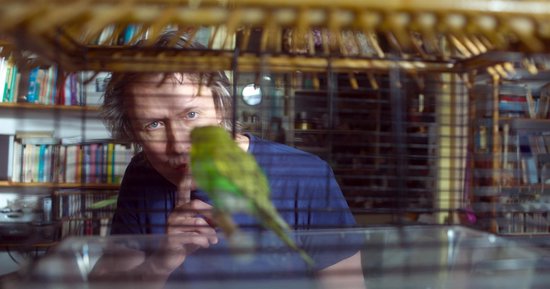 Man kijkt geheimzinnig door een vogelkooi, met een parkiet erin. Hij houdt een vinger voor zijn mond.