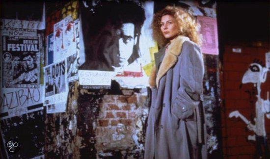 Een vrouw met een lichtgrijze trenchcoat met bontkraag staat voor een muur met posters.