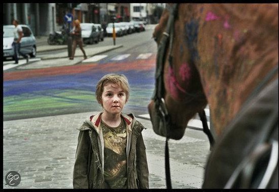 Jongen met lichtbruin haar kijkt naar de camera, met op de achtergrond een straat met gekleurde verfspatten en een paardenhoofd op de voorgrond.