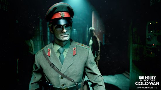 Man in militair uniform met pet en bril, staand in een donkere omgeving.