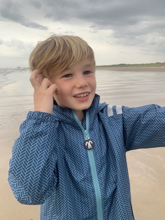 Jongen met een grijns in een blauw regenpak met visgraatpatroon, staand op het strand.