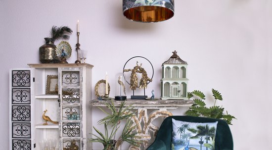 Ornements de paon dorés sur une console, avec des plumes de paon dans un vase, des bougies et une cage à oiseaux vert pâle.