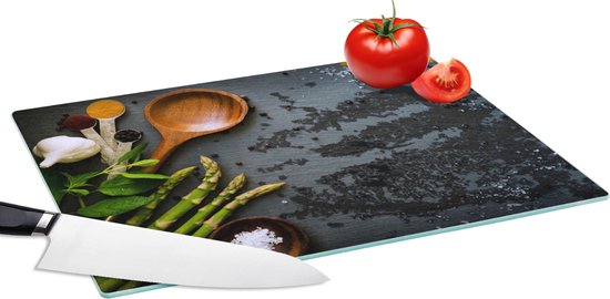 Planche à découper en verre avec texture granitée sombre, ail blanc, herbes vertes, asperges, épices dans des cuillères et tomates.