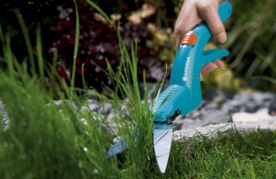 Een hand met een groen-blauwe Gardena grasschaar met oranje knop knipt gras.