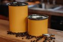 Twee matgele Airscape voorraadpotten, één groter, één kleiner, omringd door koffiebonen op een houten toonbank.