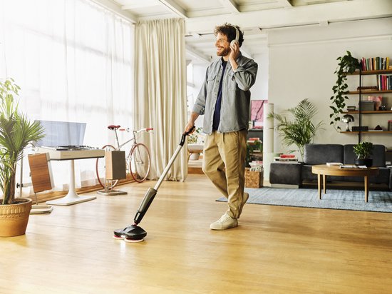 Man met koptelefoon gebruikt de zwarte elektrische vloermop in een lichte woonkamer met houten vloer.