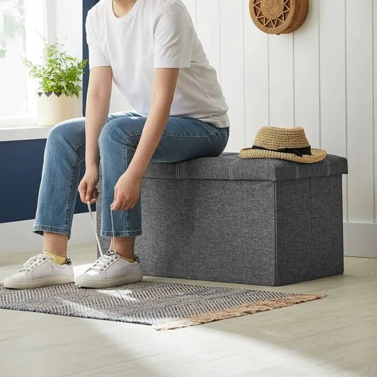 Grijze opbergkist in een hal. Een persoon in spijkerbroek en witte sneakers zit erop om schoenveters te strikken.