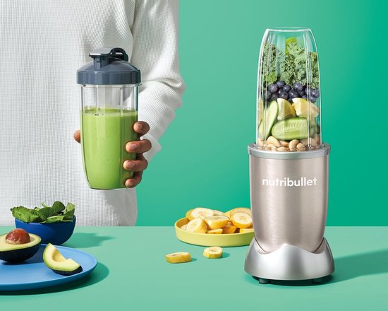 Persoon met groene smoothie in beker naast Nutribullet blender met fruit en groenten.