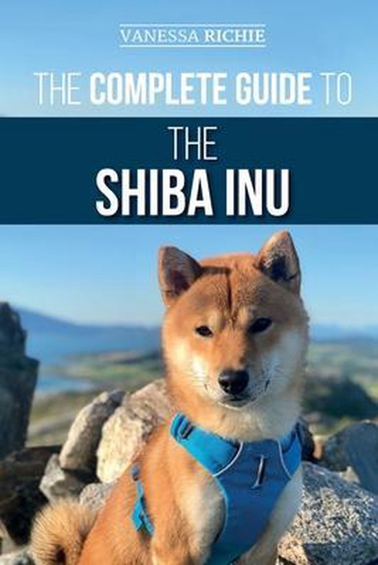 De omslag van "The Complete Guide to the Shiba Inu" toont een Shiba Inu met een blauwe halsband, zittend op rotsen met een landschap op de achtergrond.