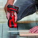 Een man in een grijze spijkerbroek knielt en gebruikt een rode en zwarte multitool met accu om hout te zagen.