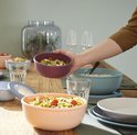 Hand houdt roze opslagbak met salade vast, op tafel staan diverse Mepal opslagbakken met eten.