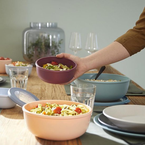 Hand houdt roze opslagbak met salade vast, op tafel staan diverse Mepal opslagbakken met eten.