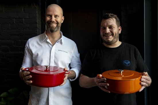 Twee mannen met baarden, een in wit hemd, de ander in zwart T-shirt, houden rode en oranje gietijzeren braadlannen vast.