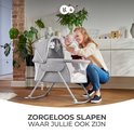 Moeder en baby glimlachen bij een grijze wieg in een woonkamer. Titel: Zorgeloos slapen.