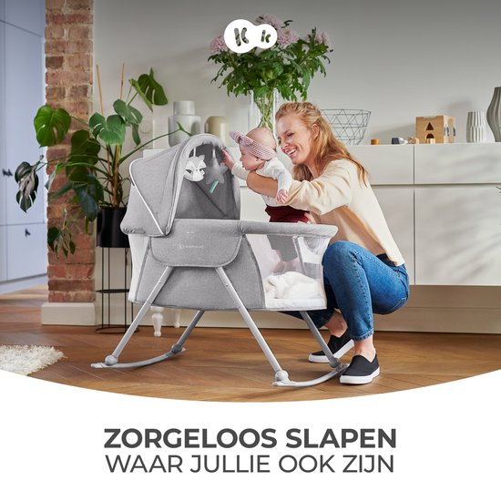 Moeder en baby glimlachen bij een grijze wieg in een woonkamer. Titel: Zorgeloos slapen.