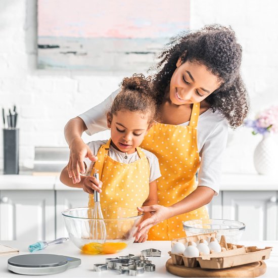 Vrouwen en meisje met gele stippen schorten in een keuken, met een keukenweegschaal op de tafel.