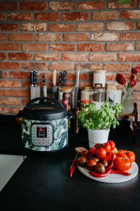 Multicooker met groen bladmotief op zwart aanrecht. Tomaten, pepers en basilicum ernaast.
