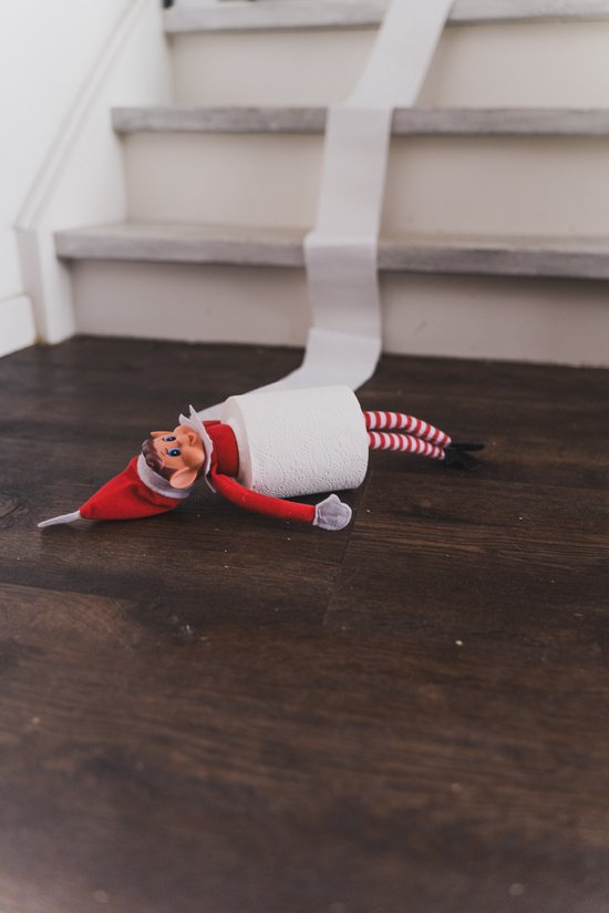 Elf met kerstmuts en gestreepte broek, verstrikt in toiletpapier op een houten vloer bij een trap.