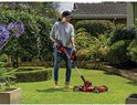 Vrouw in grijze top en spijkerbroek gebruikt rode grastrimmer met wielen in een tuin.