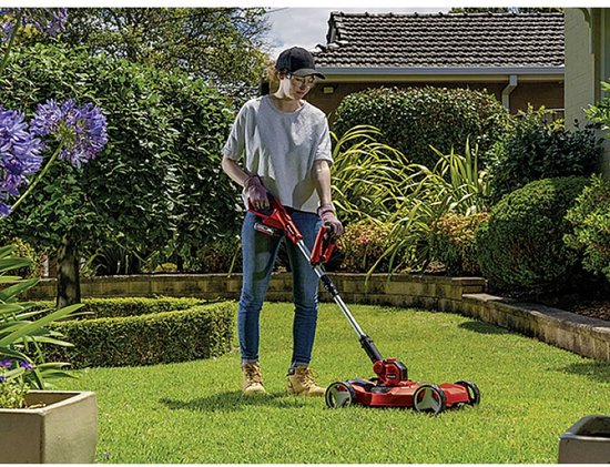 Vrouw in grijze top en spijkerbroek gebruikt rode grastrimmer met wielen in een tuin.
