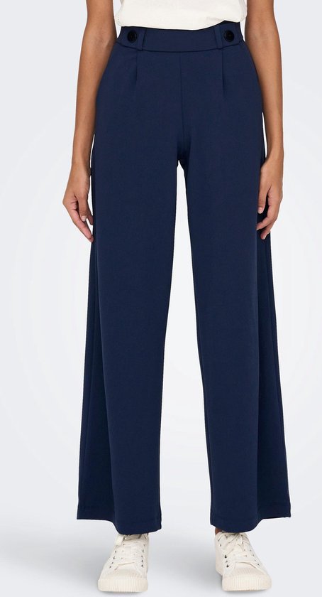 Model in donkerblauwe wijde broek met hoge taille, knopen en witte sneakers.