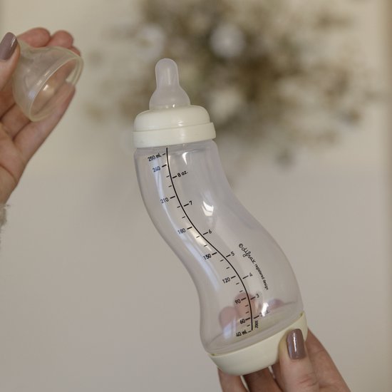 Des mains tiennent un biberon Difrax S couleur crème. Le capuchon est retiré.