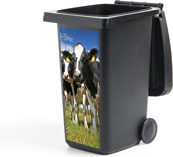 Poubelle noire avec autocollant de deux vaches noires et blanches dans un pré verdoyant sous un ciel bleu.
