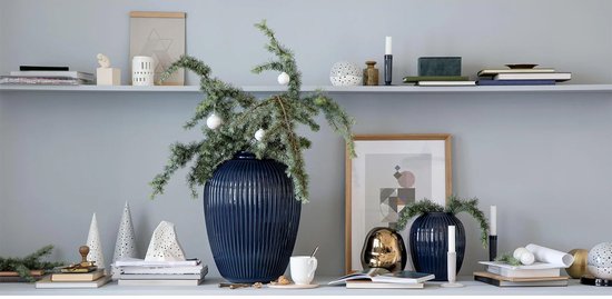 Donkerblauwe, geribbelde kandelaar op een plank bij een grote vaas met kerstgroen.