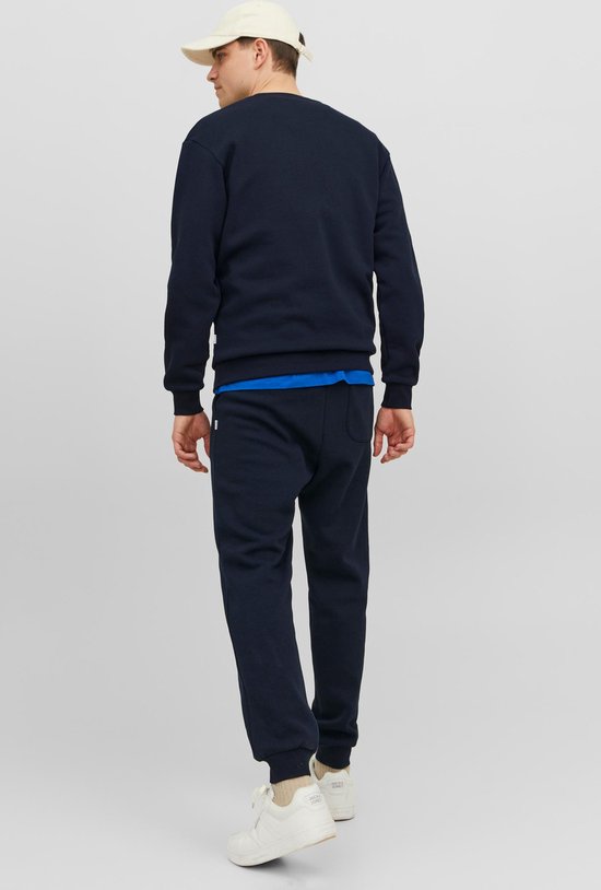 Man, achterkant, in donkerblauwe sweater en joggingbroek, met een beige pet en witte sneakers.