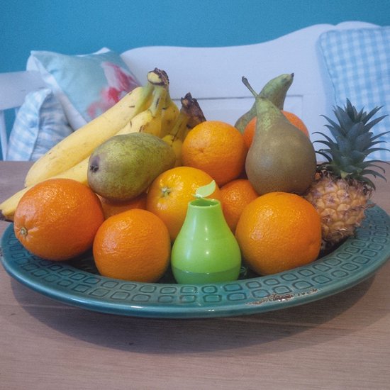 Een fruitschaal met bananen, peren, sinaasappels, ananas en een kleine groene fruitvliegjesval.