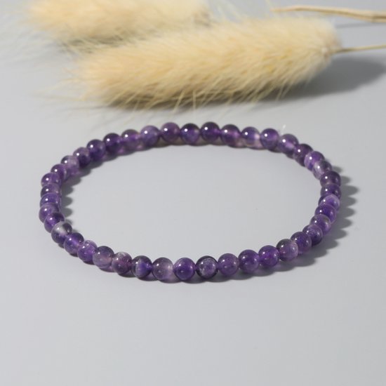 Armband van kleine, paarse amethistkralen op een grijze achtergrond met zachte pluimen.