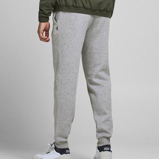 Man in een lichtgrijze joggingbroek, groene bomberjack en bruine hoodie, met witte sneakers.