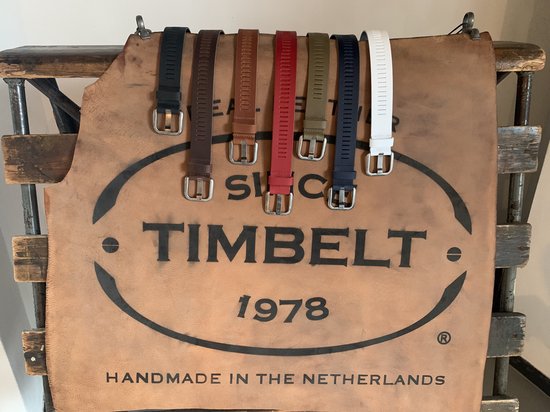 Zes TimBelt riemen in de kleuren zwart, bruin, rood, groen, marineblauw en wit, hangend over een stuk leer met TimBelt logo.