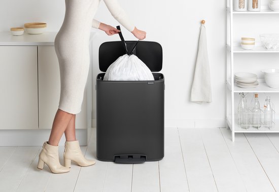 Vrouw in beige jurk en laarzen haalt een volle vuilniszak uit een donkergrijze Brabantia prullenbak.