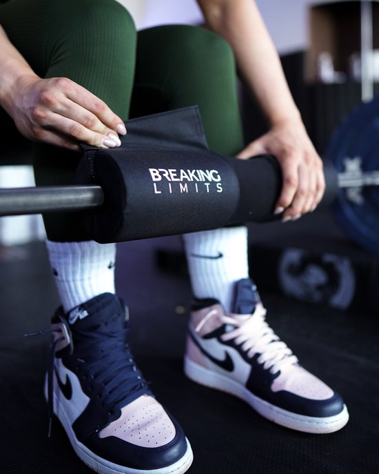 Een vrouw met groene legging en witte sokken bevestigt een zwarte halterstang pad met 'BREAKING LIMITS' opdruk.