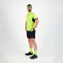 Man poseert met handen op de heupen. Draagt een fluorgeel fietsshirt, zwarte fietsbroek, groene sokken, schoenen, handschoenen en een pet.
