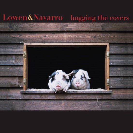 Twee varkens met zwarte vlekken op hun gezicht en oren, kijken uit een houten raam. De tekst 'Lowen & Navarro hogging the covers' staat bovenaan.