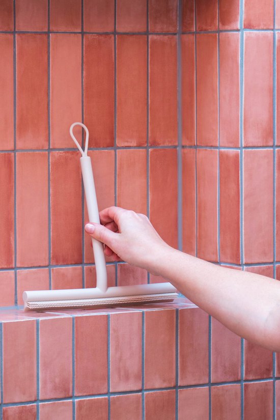 Hand met een crèmekleurige douchewisser met ophanglus tegen een achtergrond van roze tegels.