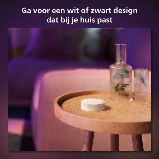 Een witte Philips Hue Tap Dial Switch staat op een houten bijzettafel in een woonkamer met paarse sfeerverlichting. De tekst zegt 'Ga voor een wit of zwart design dat bij je huis past'.