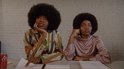 Twee personen met grote afro-kapsels zitten aan een tafel. De ene persoon draagt een gestreept shirt en de andere een paars shirt.