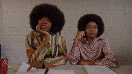 Twee personen met grote afro-kapsels zitten aan een tafel. De ene persoon draagt een gestreept shirt en de andere een paars shirt.
