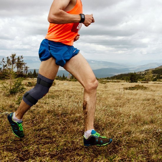 Een man in sportkleding draagt een donkergrijze kniebrace tijdens het hardlopen in een bergachtig landschap.