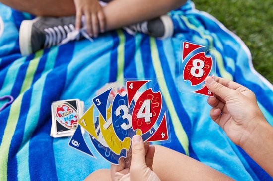 Kinderen spelen UNO H2O op een strandlaken. Een kind houdt kaarten vast met de nummers 3 en 4. Een andere kaart met nummer 8 is in de lucht.