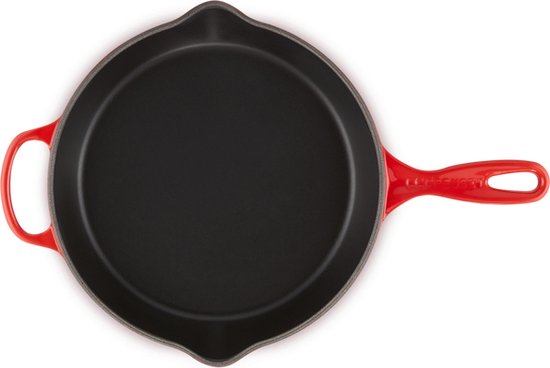 Een rode gietijzeren diepe koekenpan van Le Creuset, van bovenaf gezien. De pan heeft een zwarte binnenkant en een lange steel.