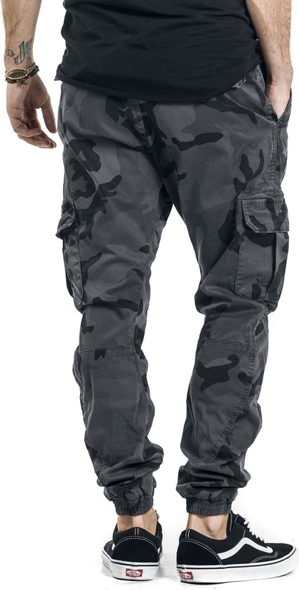 Model in donkere camouflage cargo broek met zakken en zwarte Vans sneakers.