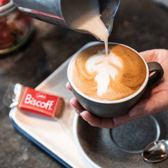 Cappuccino met melkkunst in een mok, wordt bijgeschonken, met verpakt Lotus Biscoff koekje.