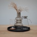 Zwart rond dienblad met een glazen karaf, glas, geribbelde vaas met siergrassen op een houten tafel.